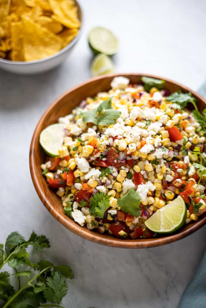 close up image of corn salad in wooden bowl alongside a bowl of chips